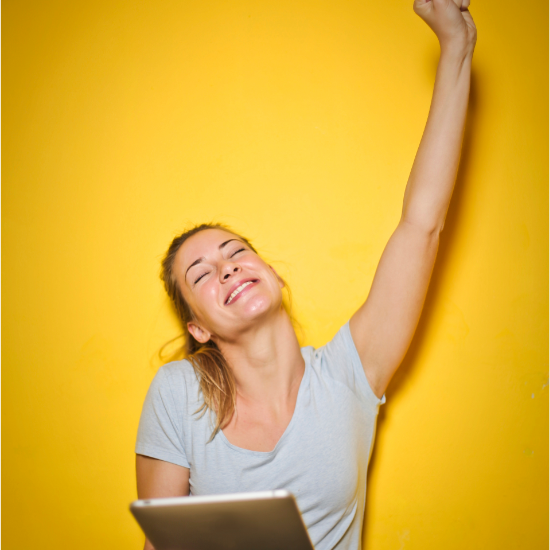 Femme souriante levant un bras, tenant une tablette, sur fond jaune.