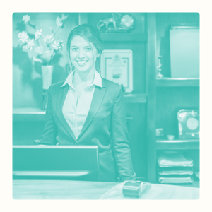 Femme souriante en costume, debout derrière un comptoir de réception. Décor chaleureux.