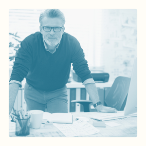Homme mature avec lunettes, se penchant sur un bureau désordonné, regardant l'objectif.